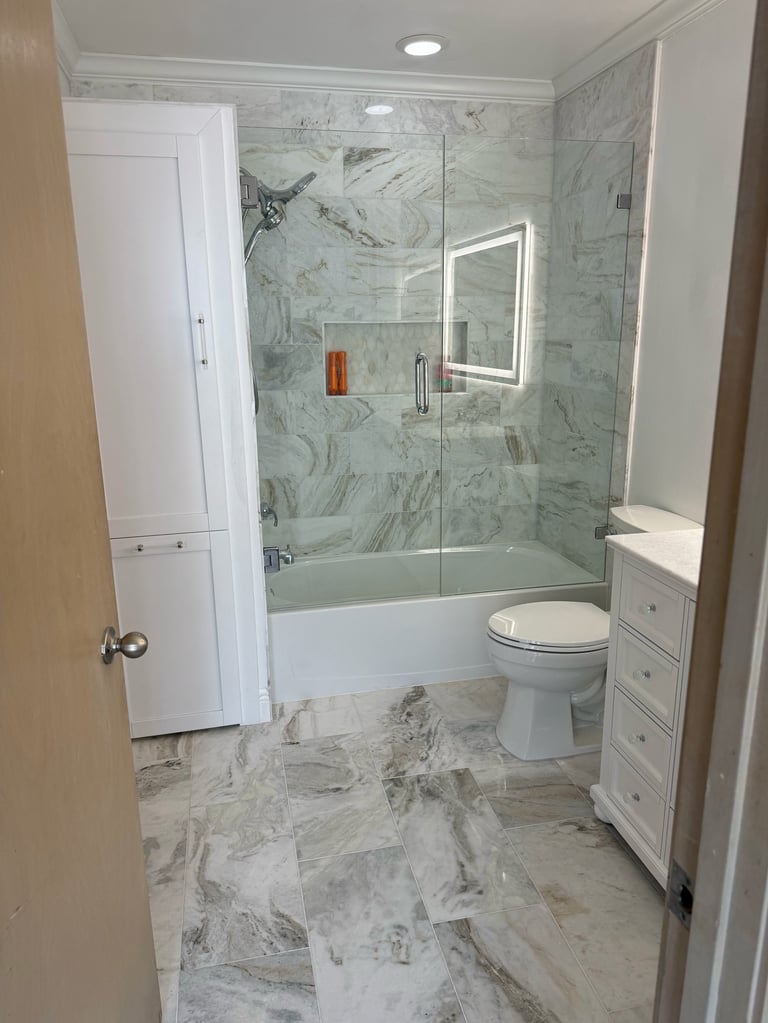 Modern bathroom with marble tile, frameless glass shower enclosure over bathtub, floating vanity, and illuminated mirror